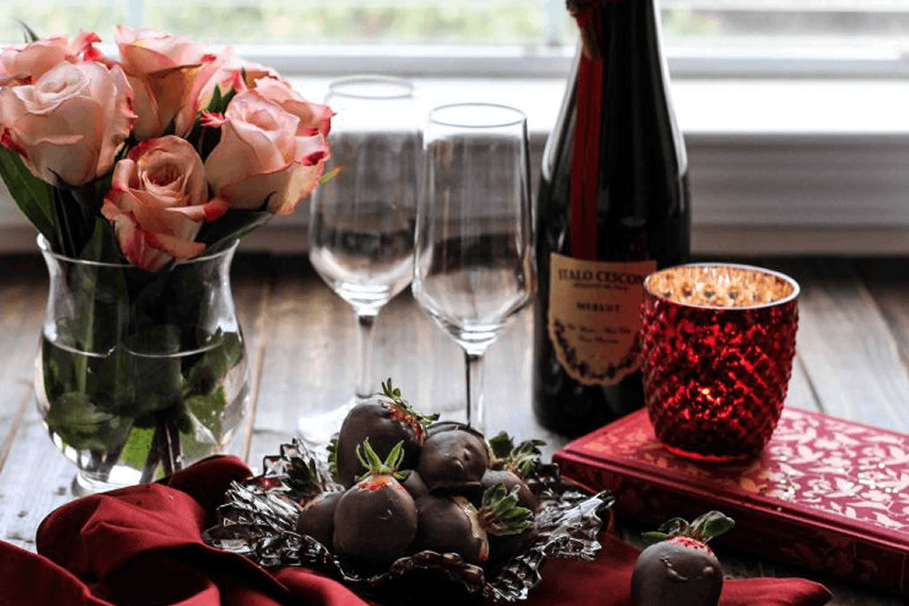 Vase of pink tinged roses, two stemmed wine glasses and chocolate covered strawberries with a votive candle in front of a window overlooking a green yard