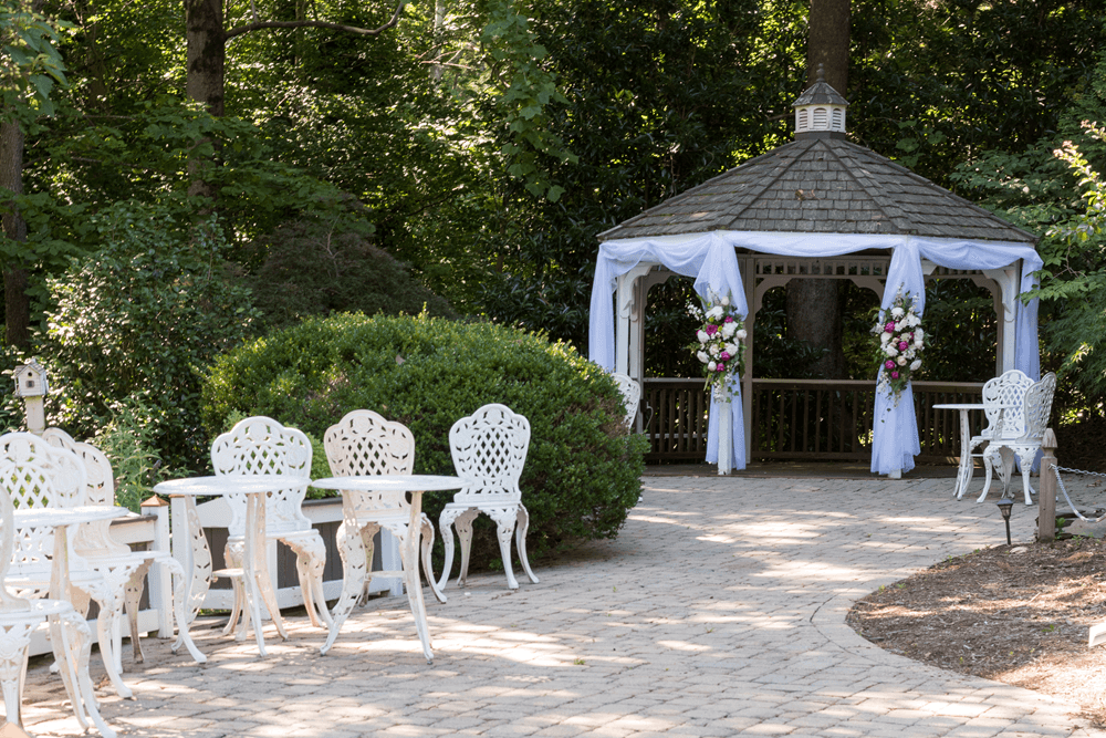 Gazebo with white drapes and flowers, iron white furniture and greenery in the background