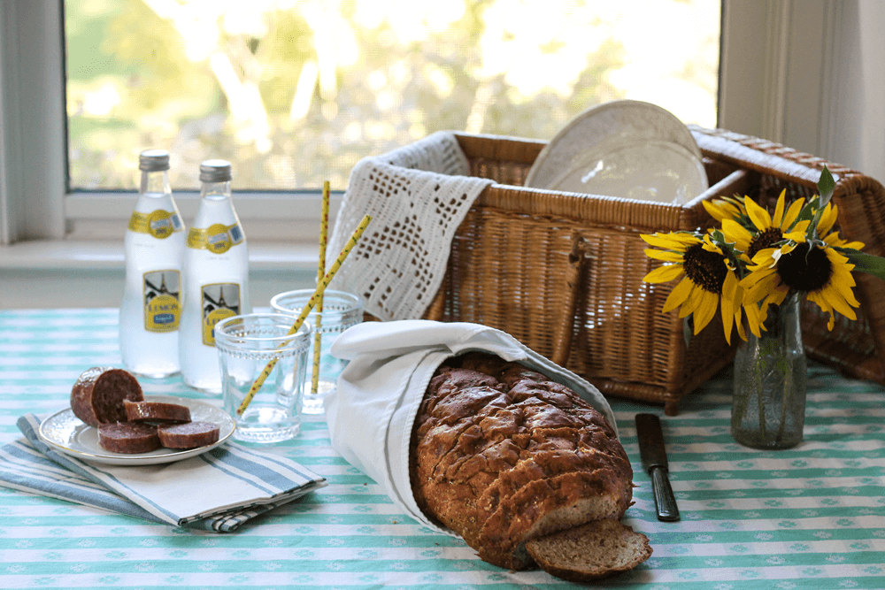 Picnic basket with plates, loaf of bread, glasses and bottled lemon water with yellow flowers in front of window overlooking yard