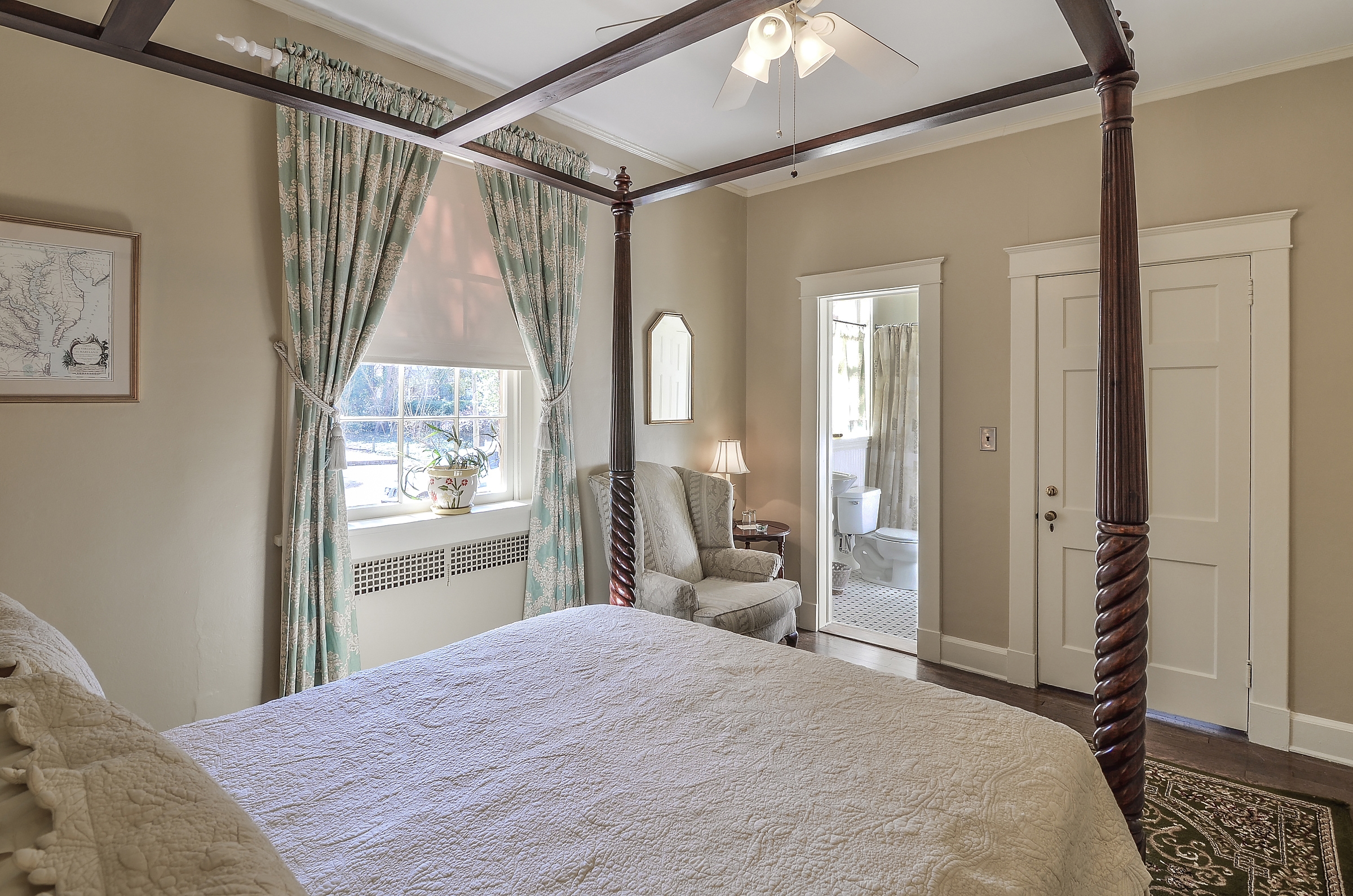 Bedroom with four-poster bed, cozy armchair, and ceiling fan and open door to the bathroom with black and white tile