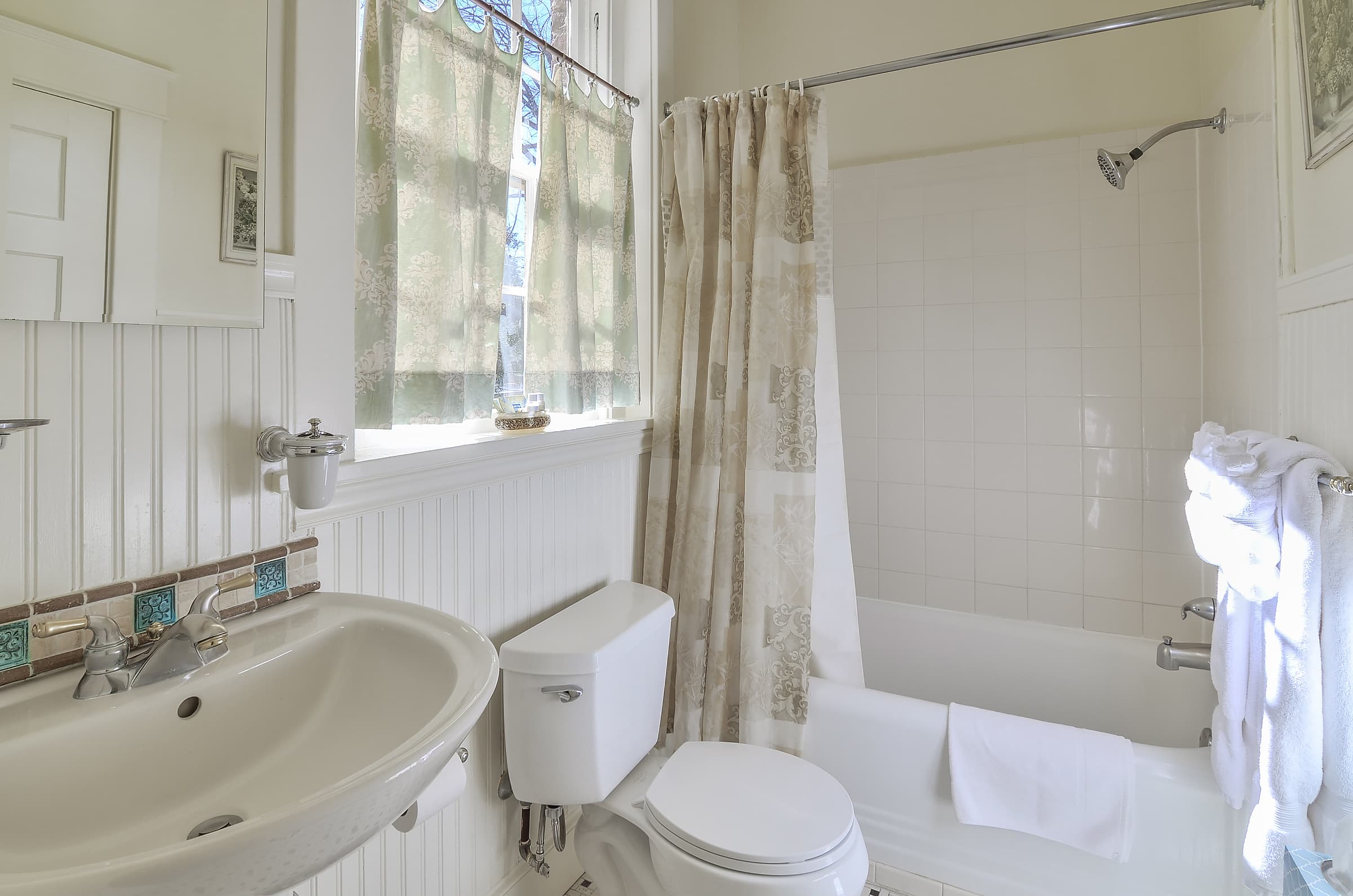 Bathroom with white wainscoting, textiles in shades of white, cream and blue, and white pedestal sink with decorative blue and tan backsplash tiles