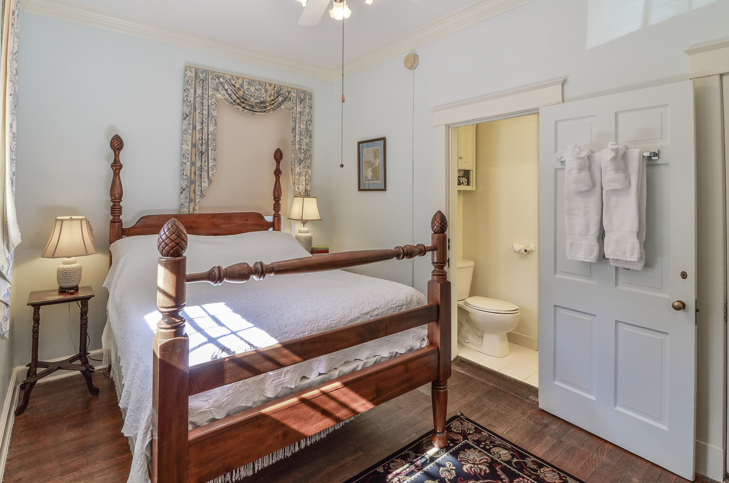 Bedroom with four-poster queen bed, oriental area rugs, nightstand with lamp, ceiling fan and open door to water closet