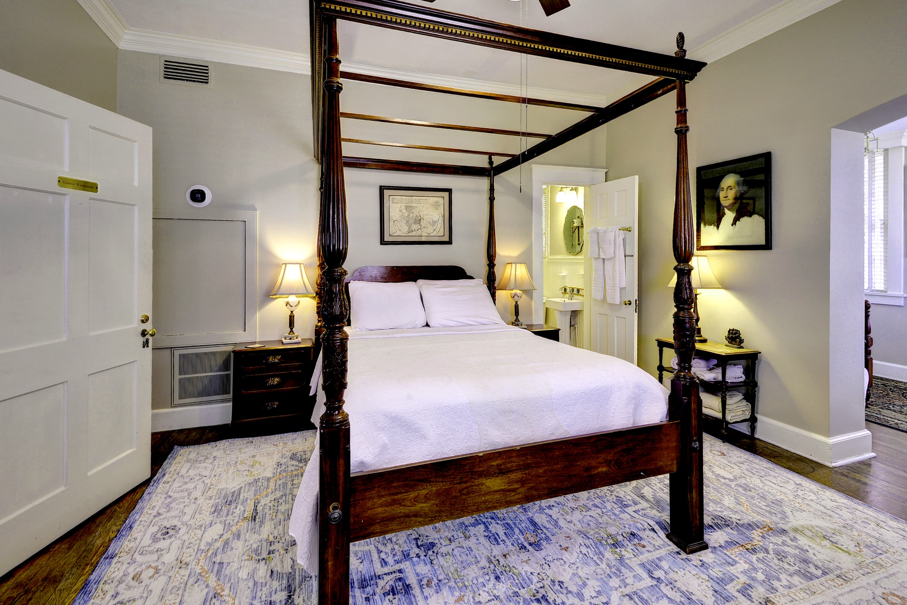 Carved mahogany four-poster bed with nightstands and lamps, hardwood floors, doorway to bathroom, and doorway leading to hallway with stairs