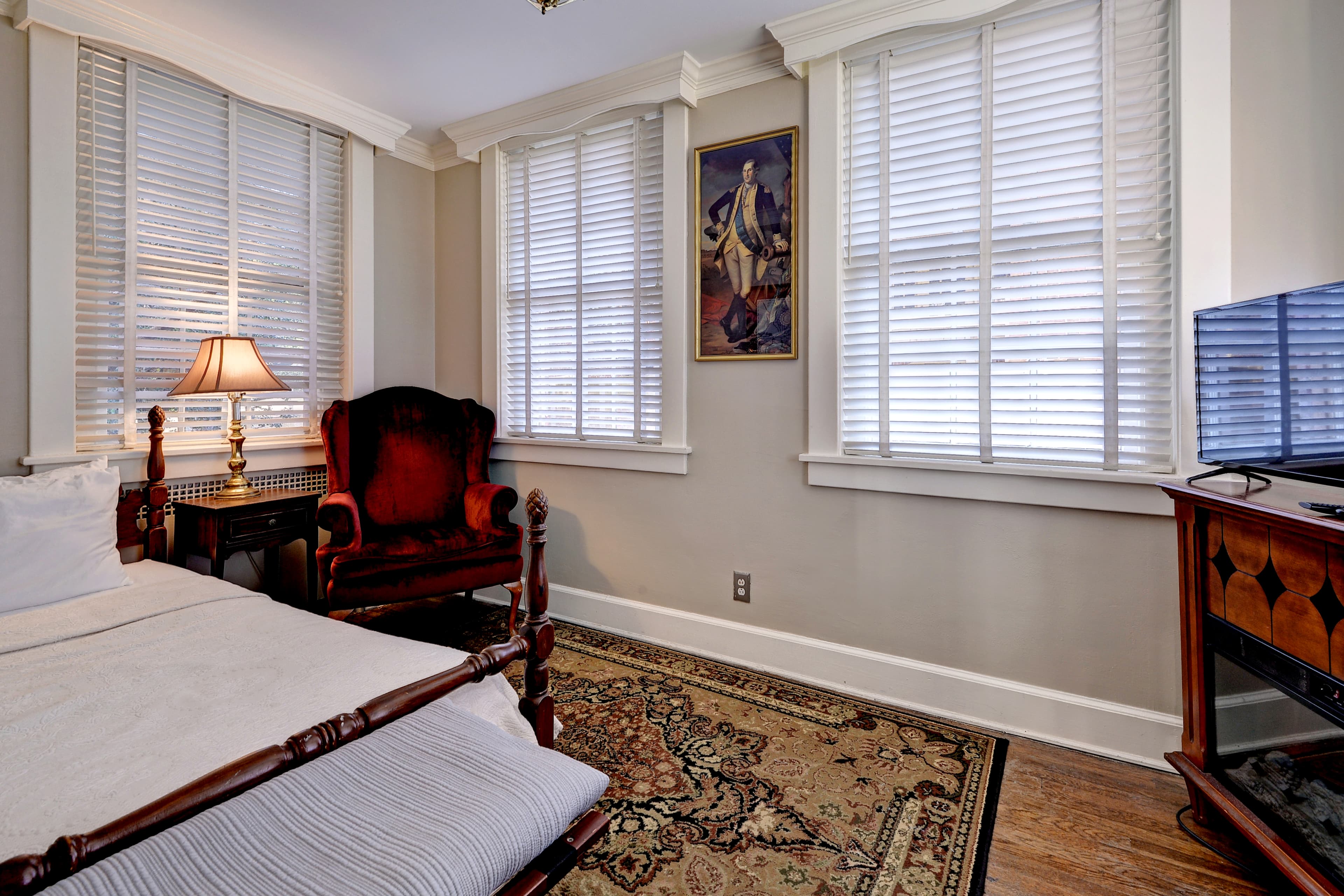 Second bedroom area with twin bed, cozy maroon armchair, bright windows and tv on wooden stand