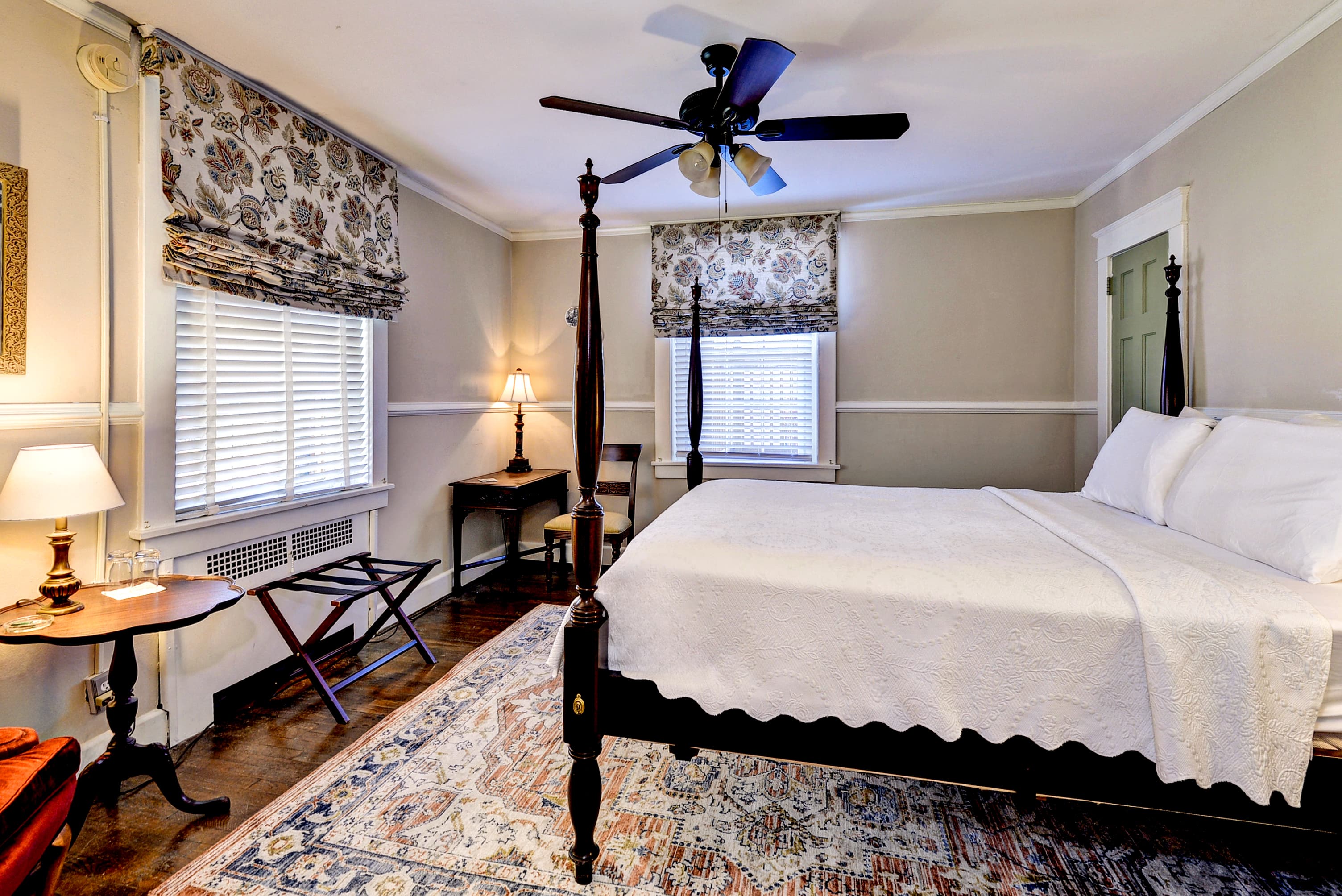 Carved mahogany four-poster bed with nightstands and lamps, hardwood floors, doorway to bathroom, and doorway leading to hallway with stairs