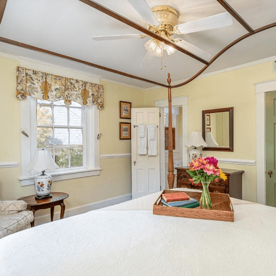 Four poster bed with white coverlet, yellow walls and wood canopy frame in bedroom with tray with flowers and books on the bed